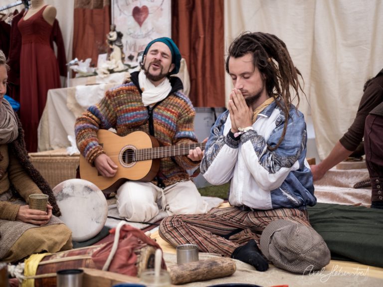 Musiker mit Gitarre und betender Mann bei intimem Folk-Konzert in rustikalem Innenraum – © Axel Hebenstreit