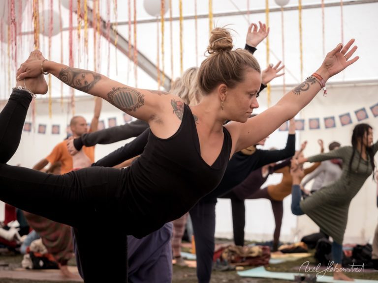 Tätowierte Yogalehrerin führt Krieger-Pose in hellem Yogastudio mit Schülern vor – © Axel Hebenstreit