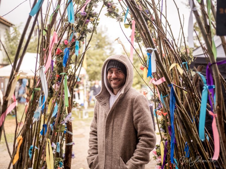 Lächelnder Mann in grauer Kapuzenjacke unter einem Bogen aus Ästen und bunten Stofffetzen auf einem Fest – © Axel Hebenstreit