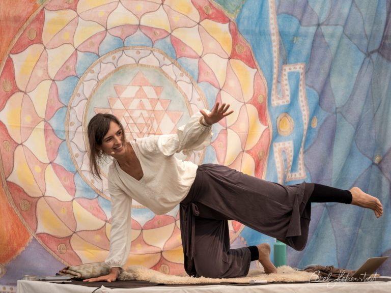Frau in weißem Oberteil und schwarzer Hose praktiziert eine spielerische Yoga-Pose vor buntem Mandala-Wandgemälde – © Axel Hebenstreit