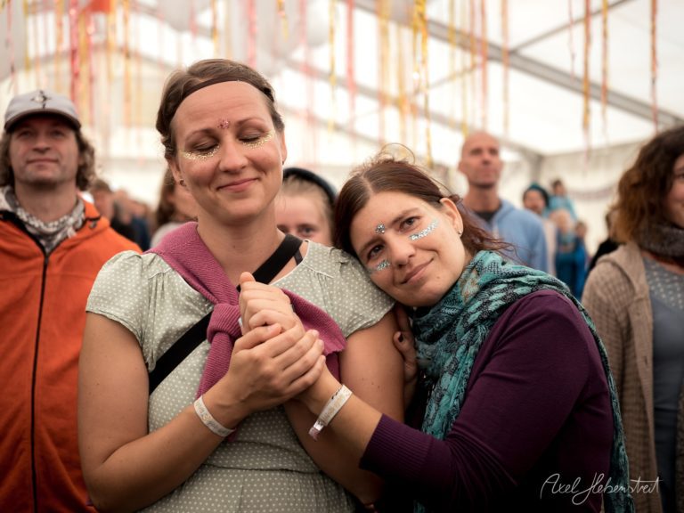 Zwei Frauen mit Glitter-Make-up umarmen sich fröhlich auf einem Festival unter Zeltdach – © Axel Hebenstreit