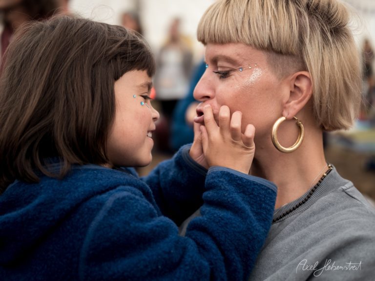 Mutter und Tochter in inniger Umarmung auf einem Festival, beide in blauer Kleidung mit emotionalem Blickkontakt – © Axel Hebenstreit
