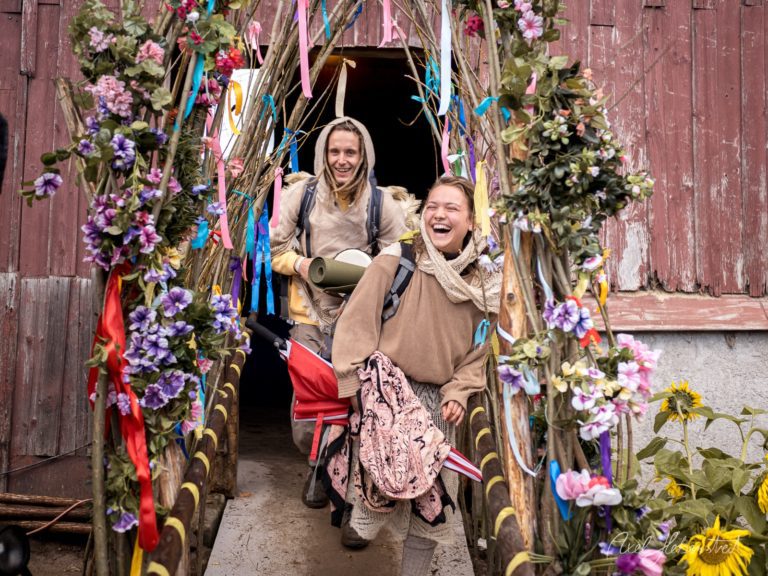 Zwei lachende Wanderinnen gehen durch ein festlich geschmücktes Tor mit bunten Blumen und Bändern – © Axel Hebenstreit