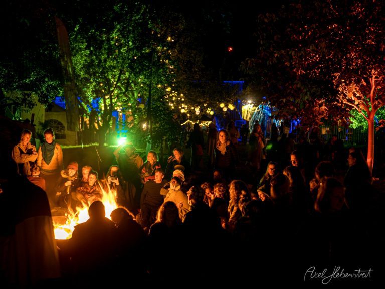 Nächtliches Lagerfeuer in einem Park mit Besuchern, die sich um das Feuer versammeln, während farbige Baumbeleuchtung die Szenerie atmosphärisch in Szene setzt – © Axel Hebenstreit