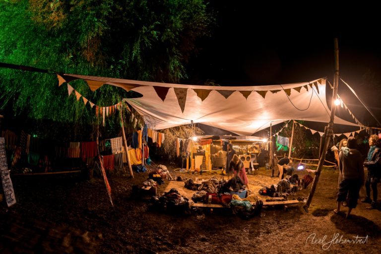 Nächtliches Festival-Lager mit beleuchteten Zelten, Wimpelketten und gesellig versammelten Besuchern um ein Lagerfeuer – © Axel Hebenstreit
