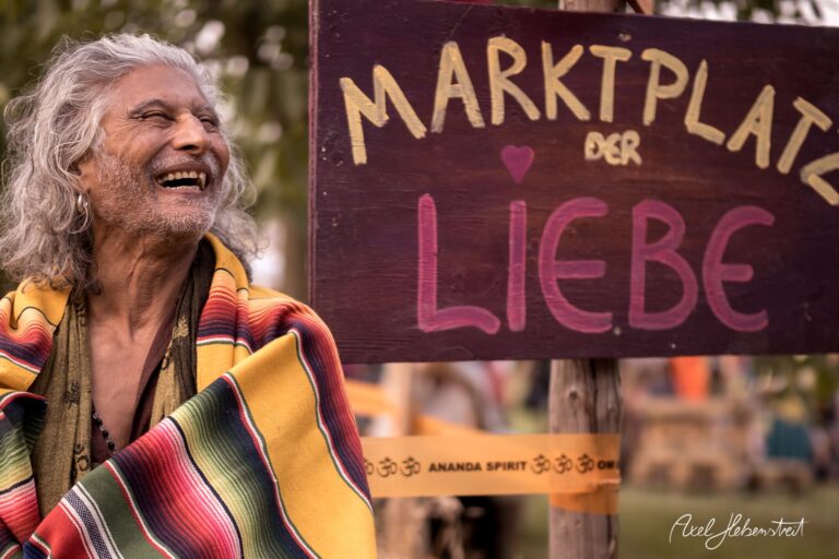 Fröhlicher Mann mit langen Haaren und bunter Decke lächelt am „Marktplatz der Liebe“ während eines Festivals – © Axel Hebenstreit