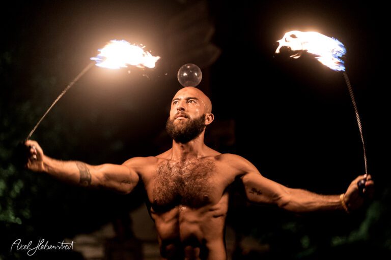 Ein athletischer Feuerartist jongliert mit brennenden Fackeln und einer Kugel auf dem Kopf, umgeben von nächtlicher Atmosphäre – © Axel Hebenstreit