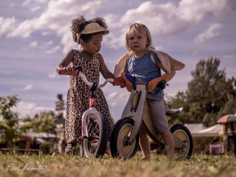 Zwei Kinder auf Spielrädern im Freien, eines in einem gepunkteten Kleid und das andere mit Flügeln, voller Abenteuerlust – © Axel Hebenstreit