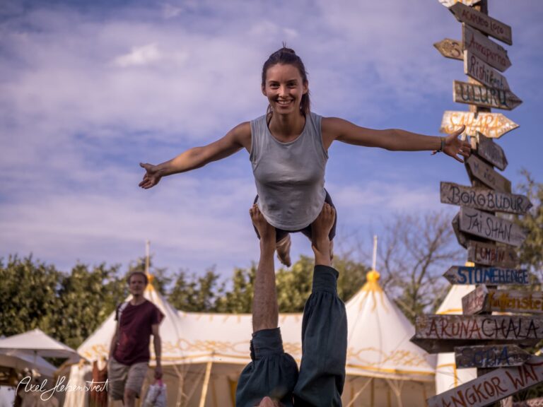 Eine schwebende Frau wird von einem Mann in einer Festivalatmosphäre gestützt, umgeben von Zelten und Orientierungsschildern. – © Axel Hebenstreit