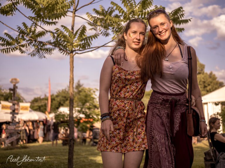 Zwei junge Frauen posieren lächelnd in einem Festival-Setting umgeben von Natur. – © Axel Hebenstreit