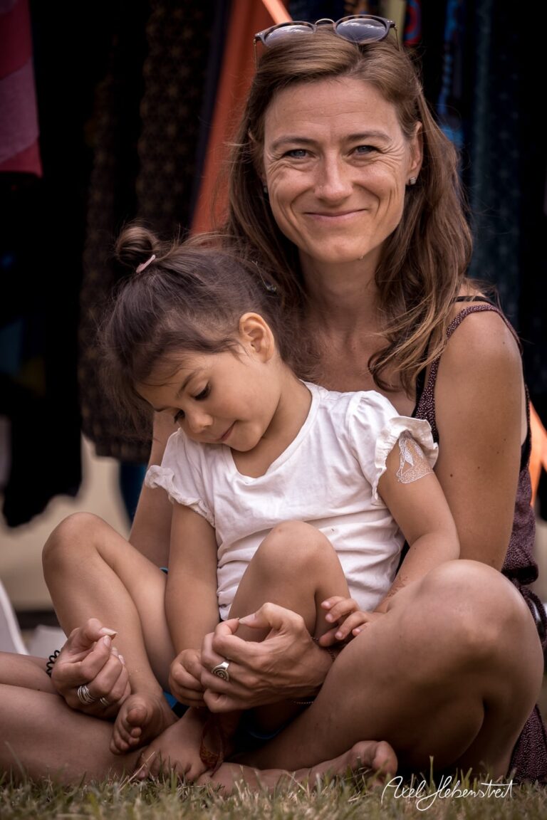 Eine lächelnde Mutter sitzt mit ihrer Tochter im Freien, während diese verträumt auf ihrem Schoß sitzt. – © Axel Hebenstreit