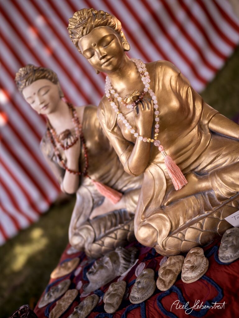 Zwei goldene Buddha-Statuen mit Schmuck, umgeben von handgearbeiteten Masken auf einem bunten Tisch – © Axel Hebenstreit
