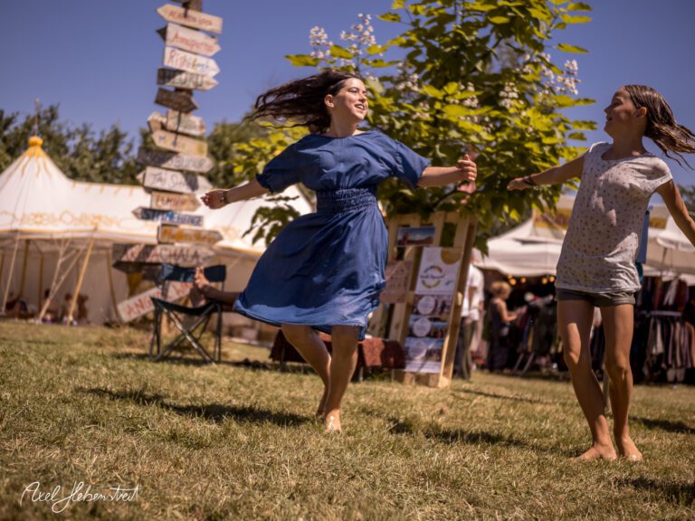 Mädchen tanzen fröhlich barfuß auf einer Wiese, umgeben von Festival-Zelt und Wegweisern – © Axel Hebenstreit