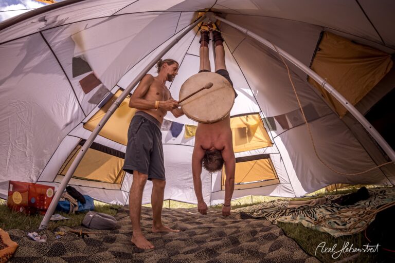 Zwei Männer in einem Zelt, einer hängt kopfüber, der andere spielt Trommel, entspannte Festivalatmosphäre. – © Axel Hebenstreit
