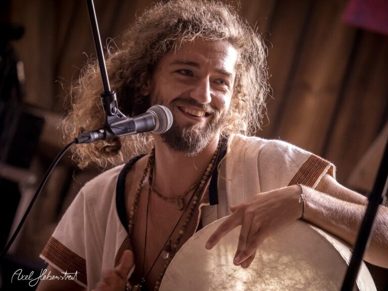 Ein lächelnder Musiker spielt ein Trommelinstrument und vermittelt eine fröhliche Stimmung auf der Bühne. – © Axel Hebenstreit