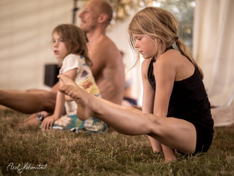 Ein Mädchen macht eine Yogaübung im Freien, während Erwachsene entspannt im Hintergrund sitzen – © Axel Hebenstreit