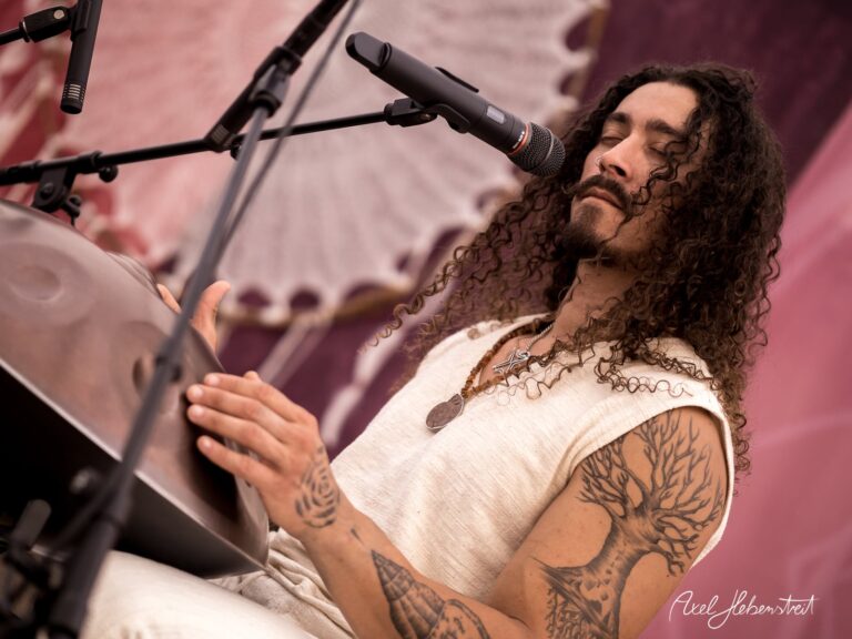 Ein Musiker mit Locken spielt mit geschlossenen Augen ein Handpan auf einer Bühne während eines Festivals – © Axel Hebenstreit