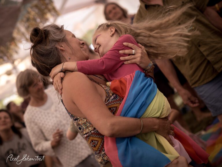 Eine fröhliche Frau tanzt mit einem Kind in bunter Kleidung auf einem Festival im Freien – © Axel Hebenstreit