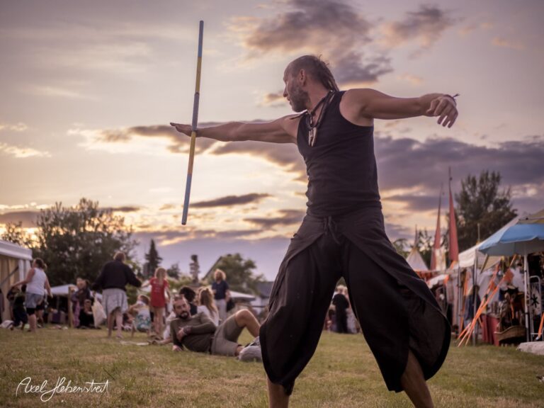 Ein Darsteller jongliert mit Stäben bei einem Festival im Freien, umgeben von einer fröhlichen Menschenmenge – © Axel Hebenstreit.