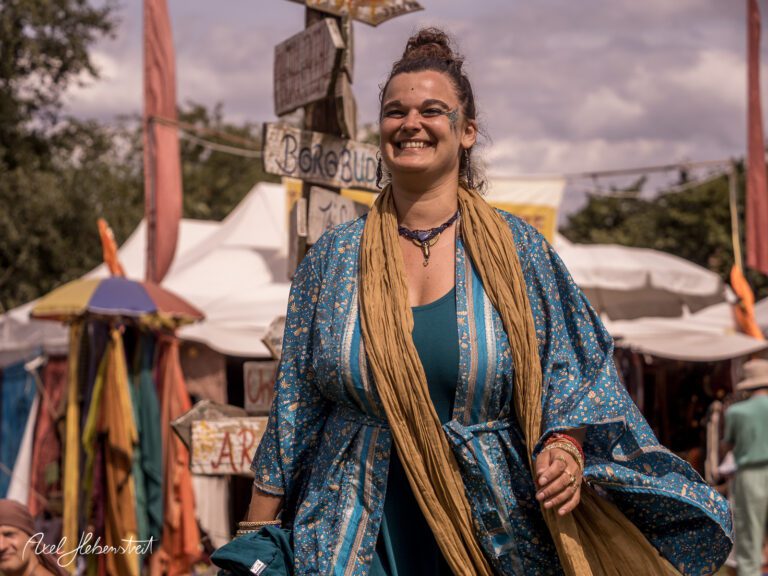 Lächelnde Frau in buntem Gewand, die fröhlich über ein Festivalgelände schlendert – © Axel Hebenstreit