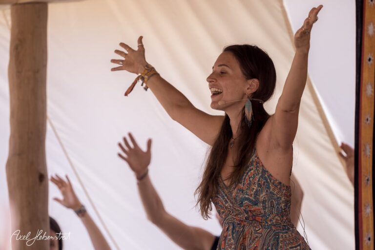 Frau in einem bunten Kleid, die fröhlich beim Yoga-Festival auf einer Bühne tanzt und anleitet – © Axel Hebenstreit
