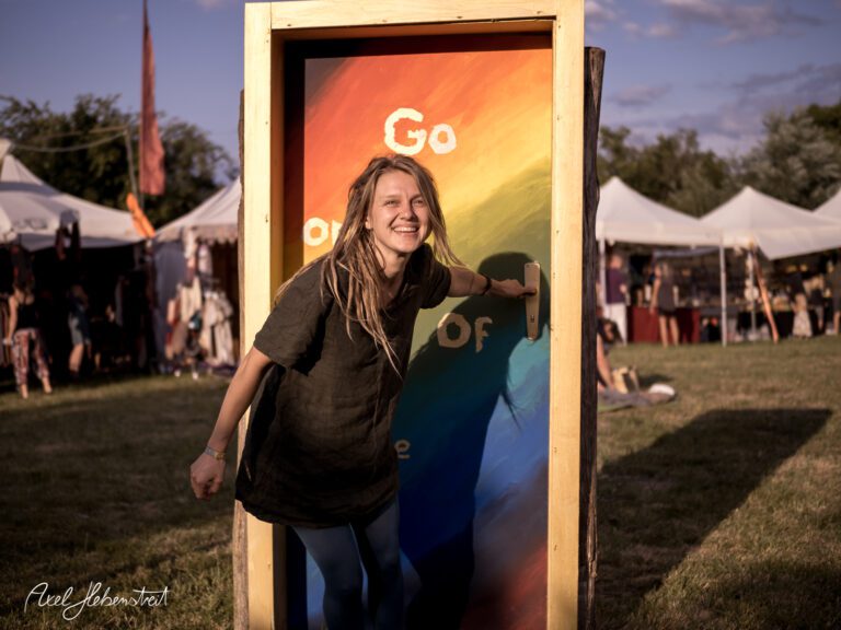 Eine junge Frau öffnet eine farbenfrohe Tür und strahlt Freude aus, im Hintergrund ein Festival-Ambiente. – © Axel Hebenstreit