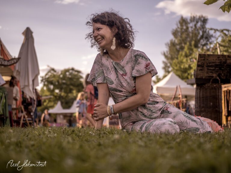 Lächelnde Frau im Pastellkleid sitzt lässig auf dem Rasen während eines Festivals im Freien – © Axel Hebenstreit