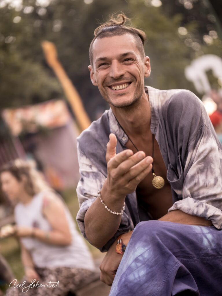 Lächelnder Mann in legerer Kleidung sitzt entspannt, umgeben von einer fröhlichen Festivalatmosphäre – © Axel Hebenstreit