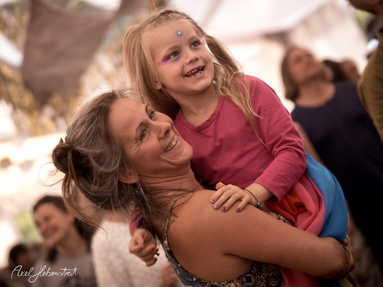 Eine fröhliche Frau hält ein lachendes Mädchen im Arm, umgeben von einer lebendigen Festivalatmosphäre. – © Axel Hebenstreit