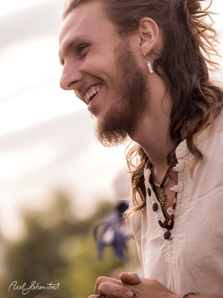 Junger Mann mit langen Haaren lächelt glücklich während eines Festivals im Freien – © Axel Hebenstreit
