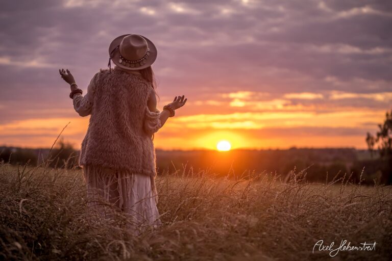 Frau in einem fließenden Kleid und Hut steht umarmend in einer Wiese bei Sonnenuntergang – © Axel Hebenstreit