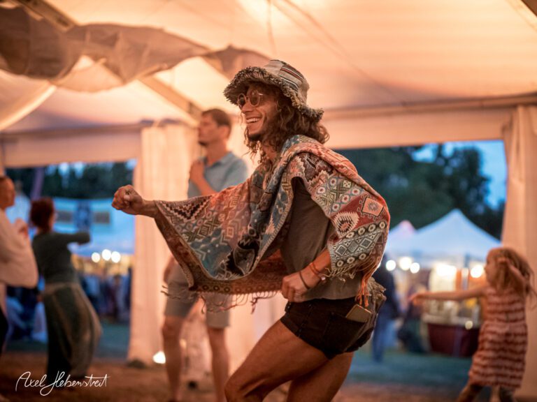 Ein fröhlicher Tänzer in Boho-Kleidung genießt die Musik auf einem Festival in einem Zelt – © Axel Hebenstreit