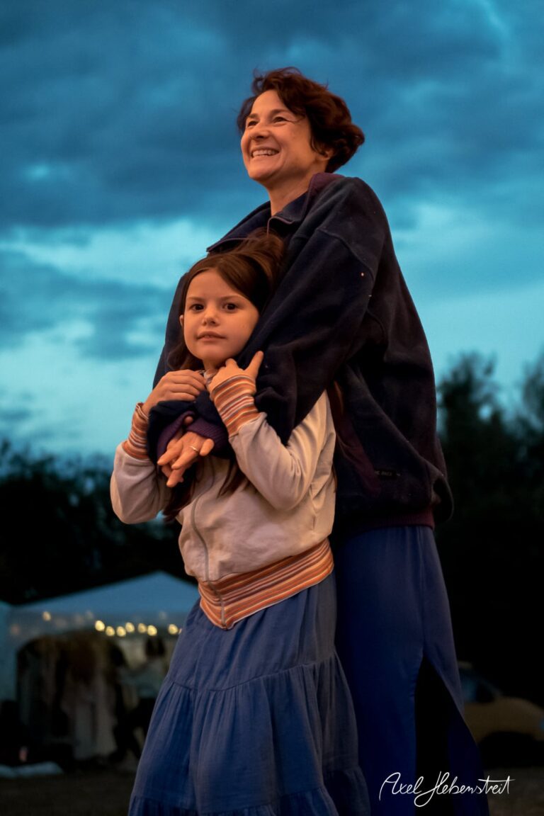 Eine lächelnde Frau umarmt ein Kind, während der Abendhimmel in sanften Farben leuchtet. – © Axel Hebenstreit
