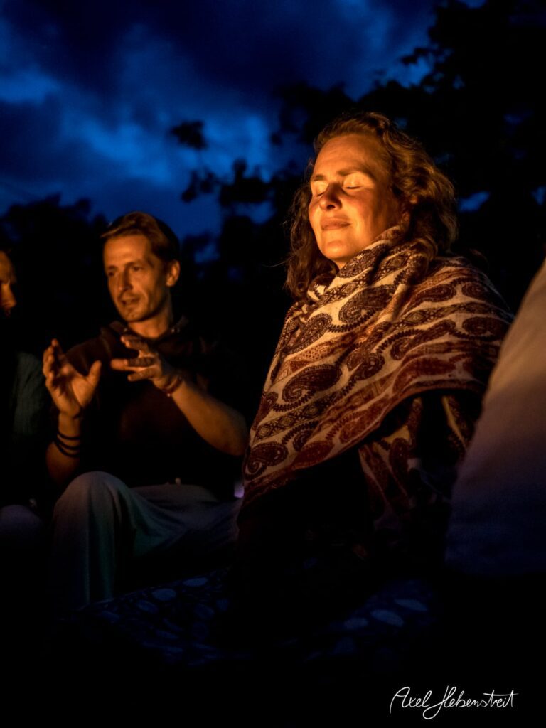 Eine nachdenkliche Frau in einem Tuch, umgeben von Gleichgesinnten am Lagerfeuer in der Dämmerung – © Axel Hebenstreit