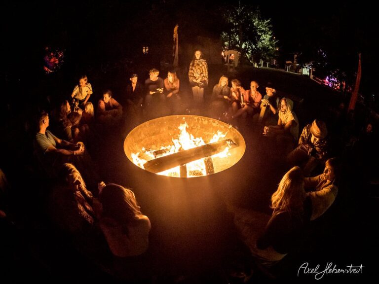 Gemütliche Lagerfeuer-Runde mit Menschen in nächtlicher Stimmung in einem Wald, umgeben von Bäumen – © Axel Hebenstreit