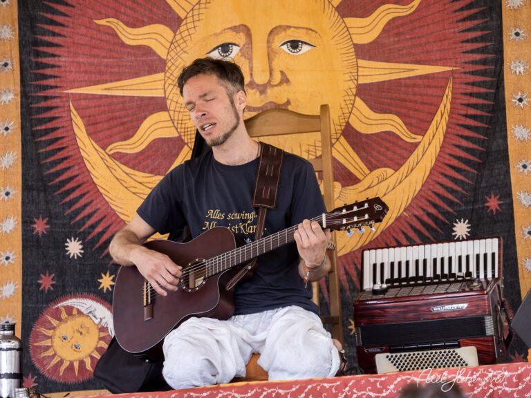 Musiker mit Gitarre singt leidenschaftlich vor einem sonnigen Wandteppich auf einer Festivalbühne – © Axel Hebenstreit