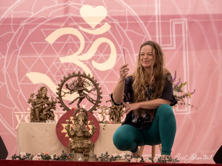Eine lächelnde Frau im Yogastil sitzt vor dekorativen Statuen auf einer Festivalbühne – © Axel Hebenstreit