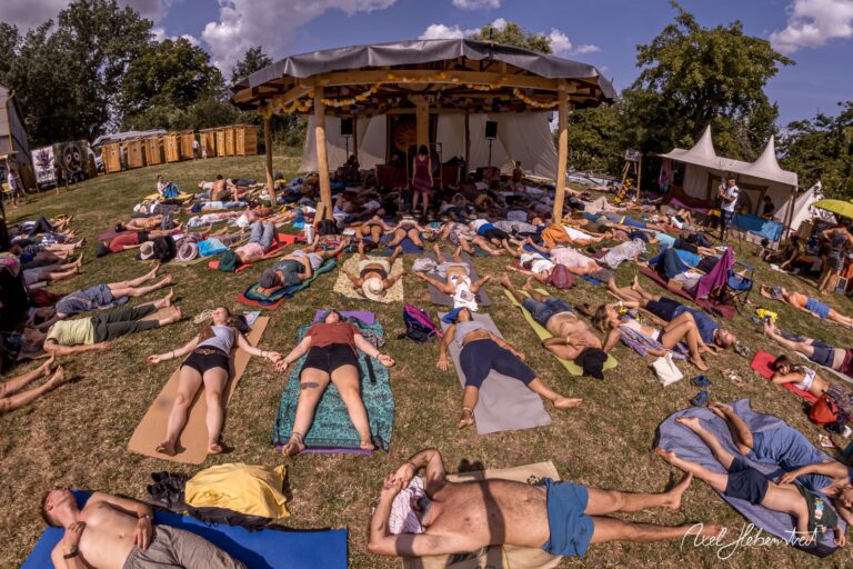 People practice yoga auf matten in der Natur bei einem Festival, umgeben von Bäumen und einem großen Zelt – © Axel Hebenstreit