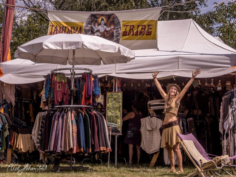 Eine fröhliche Frau mit erhobenen Armen steht vor einem bunten Verkaufsstand auf einem Festival. – © Axel Hebenstreit