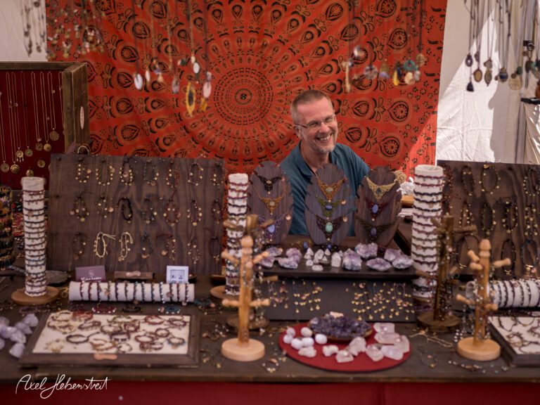Lebhafter Markstand mit einem freundlichen Verkäufer, umgeben von handgefertigtem Schmuck und farbenfrohem Hintergrund – © Axel Hebenstreit