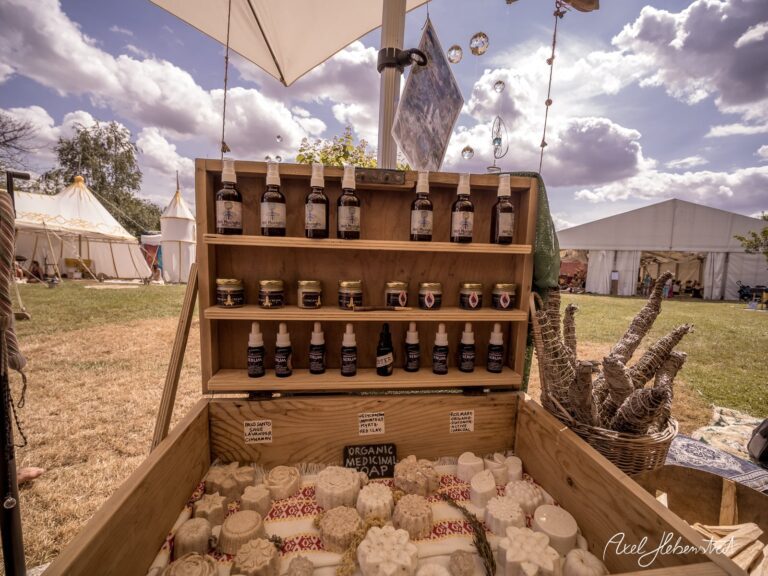Holztisch mit verschiedenen Fläschchen und Seifen auf einem Festivalgelände unter bewölktem Himmel – © Axel Hebenstreit