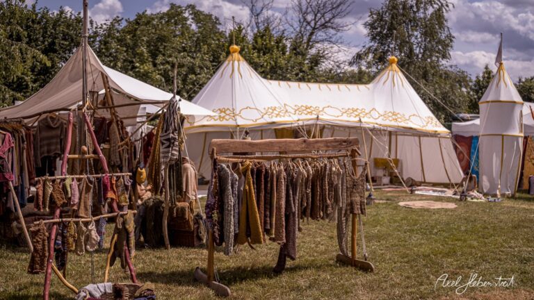 Bunte Stoffe hängen an einem Gestell vor festlichen Zelten auf einem lebendigen Festivalgelände – © Axel Hebenstreit.