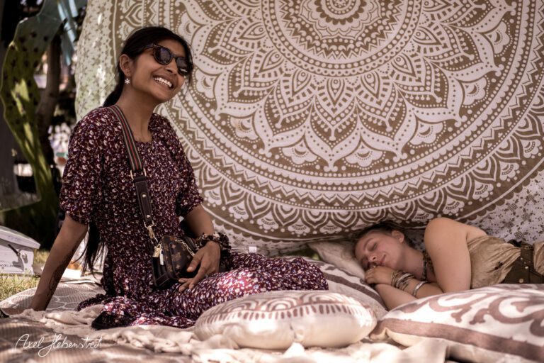 Zwei Frauen entspannen auf weichen Kissen unter einem bunten Wandbehang während eines Festivals – © Axel Hebenstreit.