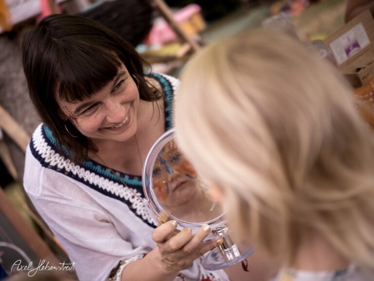 Eine lächelnde Frau zeigt einem Kind im Spiegel bunte Gesichtsmalerei auf einem Festival. – © Axel Hebenstreit