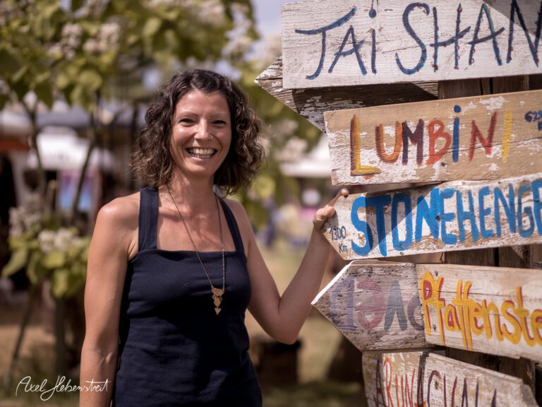 Lächelnde Frau steht neben einem bunten Wegweiser auf einem Festivalgelände – © Axel Hebenstreit