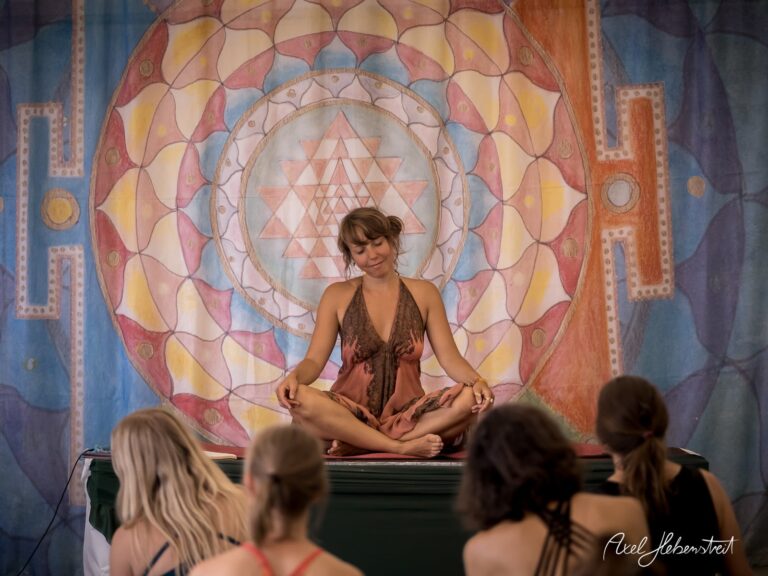 Eine Yoga-Lehrerin meditiert entspannt vor einer bunten Wandmalerei, während Zuschauer gebannt zuhören. – © Axel Hebenstreit