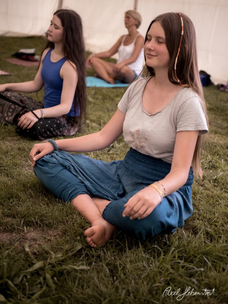 Drei Frauen meditieren entspannt im Gras während eines Yoga-Events in einem Zelt. – © Axel Hebenstreit