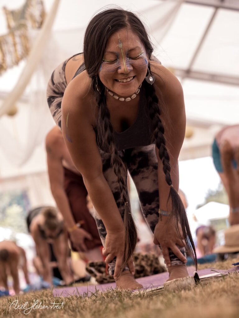 Eine Yoga-Praktizierende mit Zöpfen und Körperbemalung übt eine Vorwärtsbeuge auf einem Festivalgelände. – © Axel Hebenstreit
