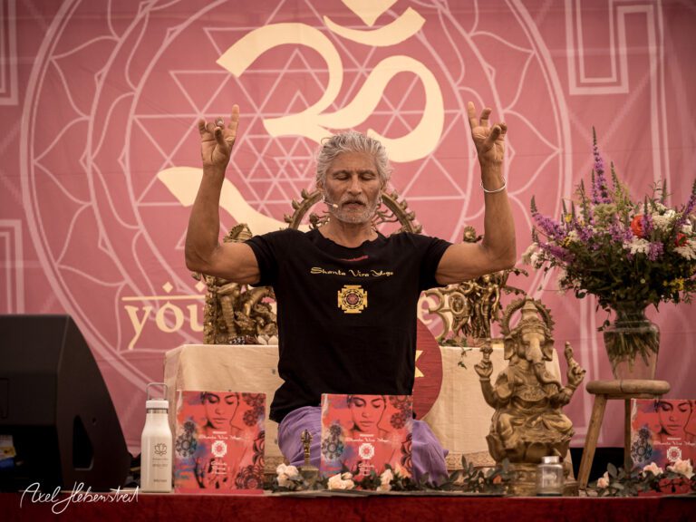 Ein yogischer Lehrer sitzt meditiert vor einer dekorativen Kulisse und erhebt die Hände in einer spirituellen Geste – © Axel Hebenstreit