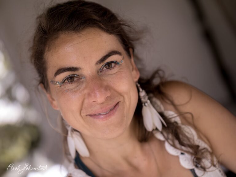 Porträt einer lächelnden Frau mit strahlendem Make-up und auffälligem Schmuck, die positive Energie ausstrahlt – © Axel Hebenstreit